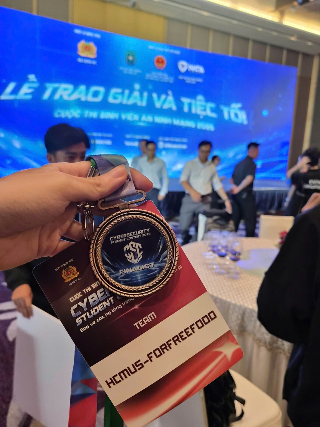 A hand holding the bronze Cybersecurity Student Contest 2026 finalist medal and the team lanyard card reading HCMUS-ForFreeFood, at the award ceremony in front of the lit stage backdrop