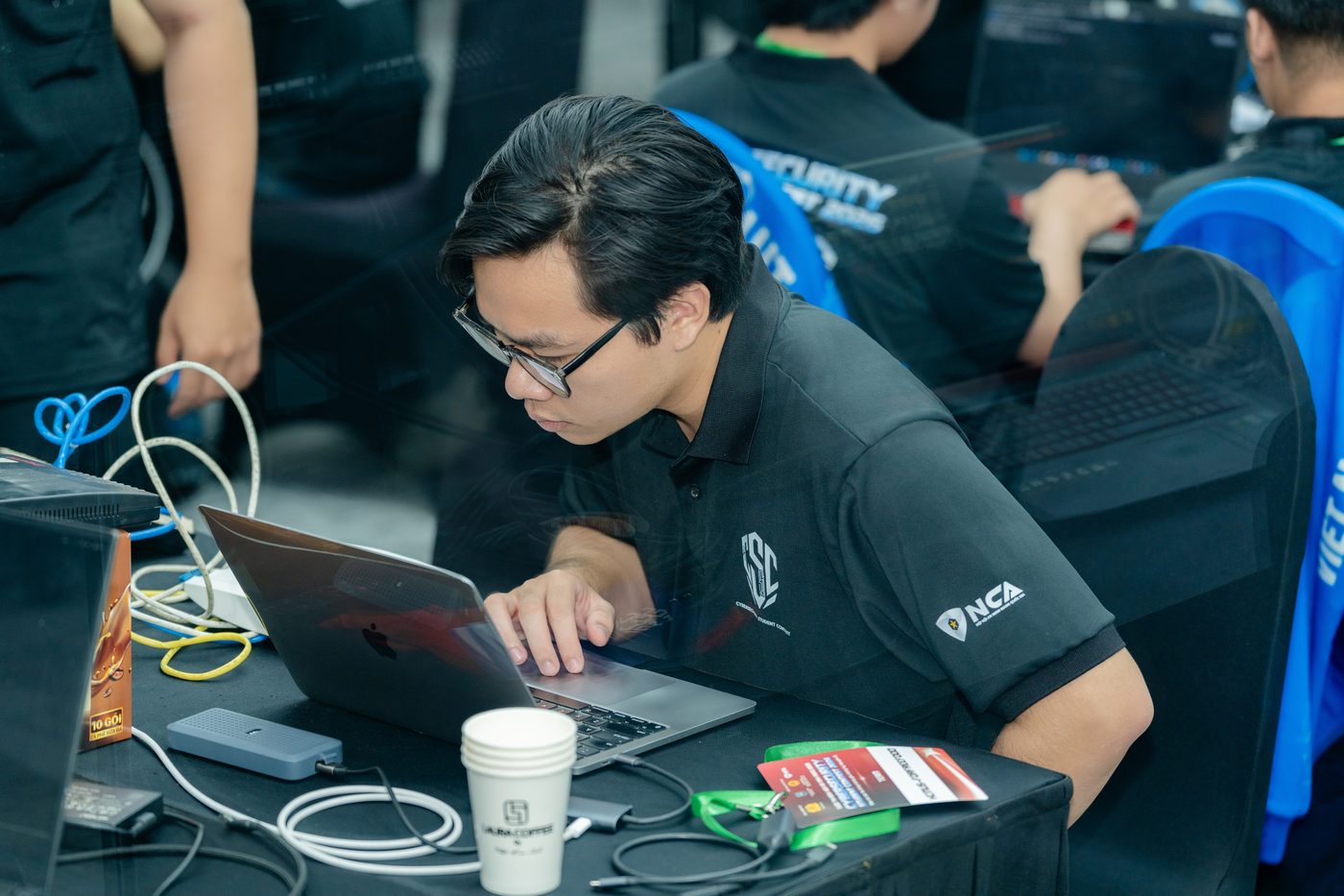 Working on a laptop during the Cybersecurity Student Contest 2026 final round, in the contest polo shirt, at a table of networked machines