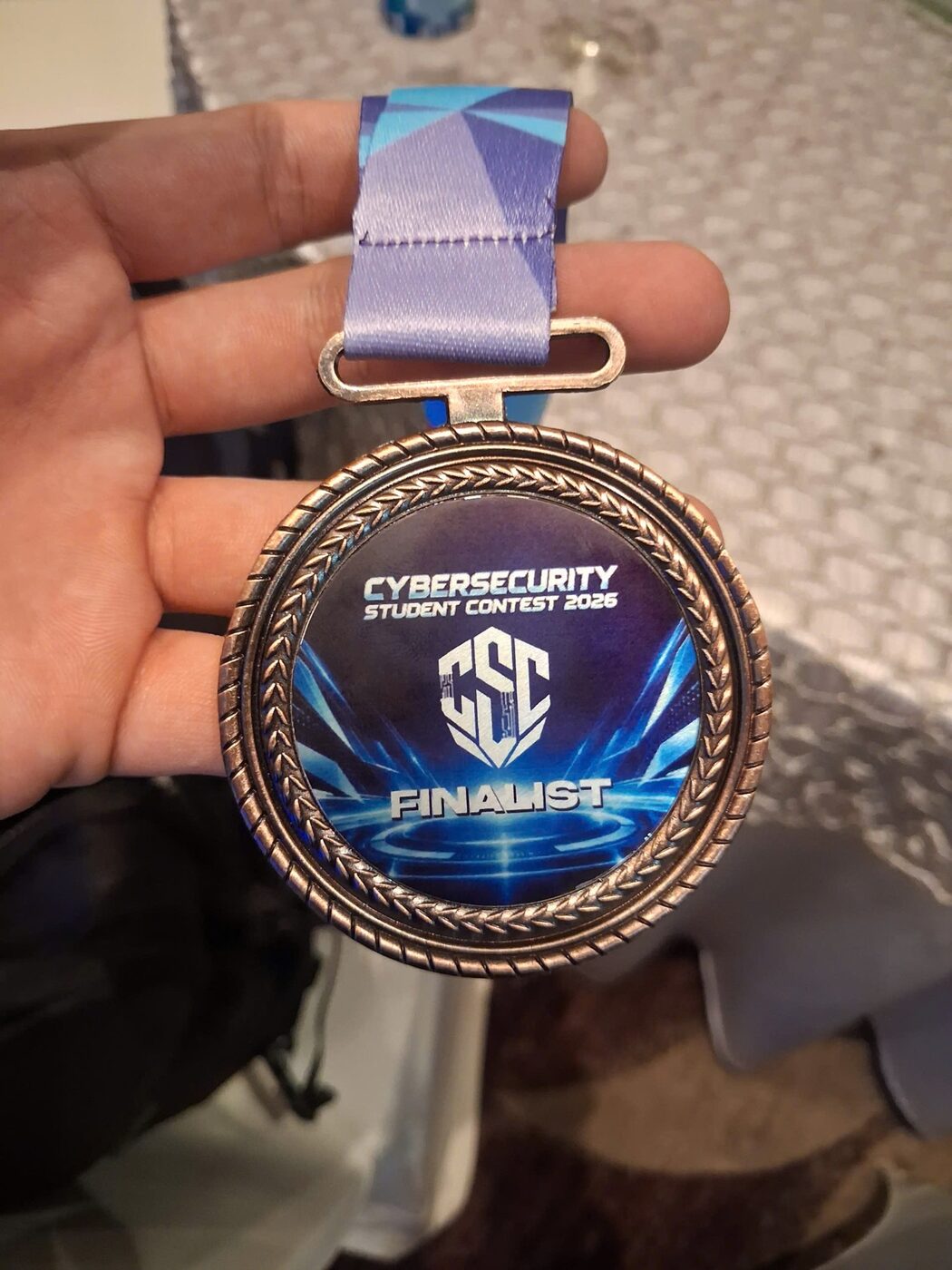 The bronze Cybersecurity Student Contest 2026 finalist medal held in one hand, its enamel face reading CSC Finalist